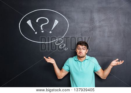 Confused perplexed young man shrugging shoulders over chalkboard background with blank speech bubble