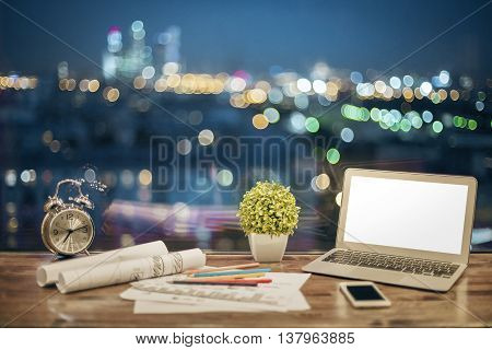 Front view of creative designer desktop with blank white laptop stationery items plant alarm clock and smartphone on blurry night city background. Mock up