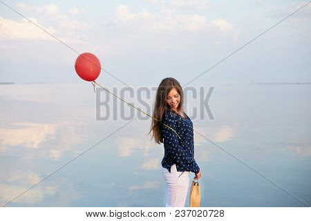 Portrait Of Young Woman With Red Air Balloon And Present Bag Near The Calm Sea Or Lake. Clouds Are R