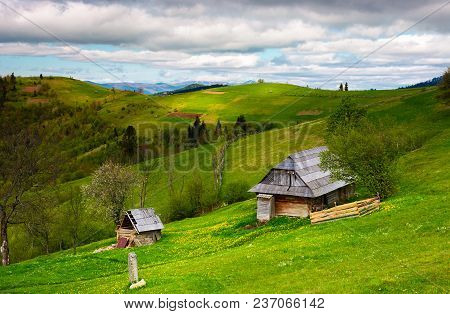 Woodshed On A Grassy Hillside On A Cloudy Day. Village Outskirts With Rural Fields In Mountainous Ar