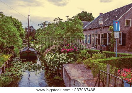 Drimmelen, Southern Netherlands - June 30, 2017. Canal With Boat Next To A Brick Rustic House And Fl