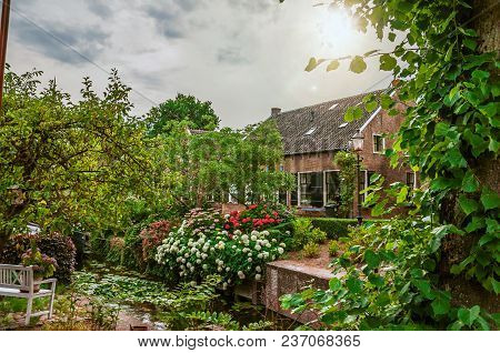 Charming Small Canal Next To A Brick Rustic House And Lush Flowery Garden In Cloudy Day At Drimmelen