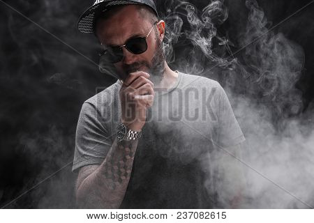 Pensive Black Bearded Male Dressed In A Grey Shirt, Sunglasses And Baseball Cap Vaping. Man In Holdi