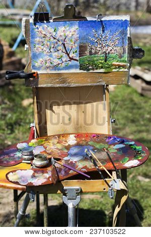 Plein Air Etude With Two Paintings And A Palette. A Palette With Multicolored Paints And Brushes Lie