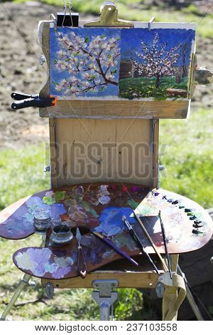 Plein Air Etude With Two Paintings And A Palette. A Palette With Multicolored Paints And Brushes Lie