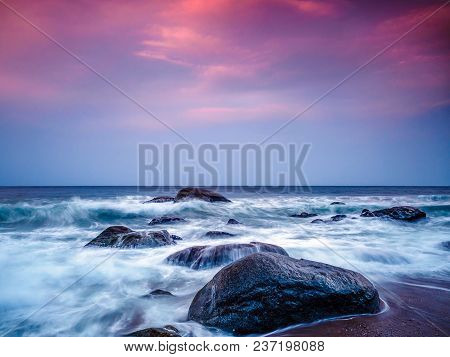 The Tide Rushes In On Some Rocks On The Beach As The Sunset Lights Up Some Clouds In The Sky, Off Th