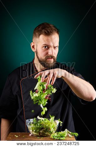 Portrait Of A Handsome Mischevious Male Chef Cook In Black Uniform Making Delicious Vegetable Salad 