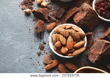 Dark Chocolate Bar Broken In Pieces On Dark Background, Cocoa Powder, Various Nuts And Raisins In Bo