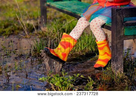 Faceless Shot Of Little Girl In Vivid Skirt And Rain Boots Swinging With Legs In Puddle Sitting On B