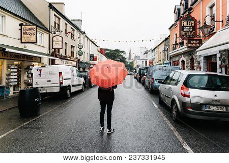 Kenmare, Ireland - November 10, 2017: Woman With Red Umbrella In The Middle Of The Street With Pictu