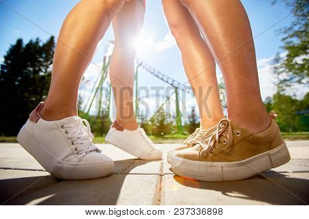 Tanned slim legs of two girls in white and beige leather cross-shoes standing in front of one another in theme park on sunny day