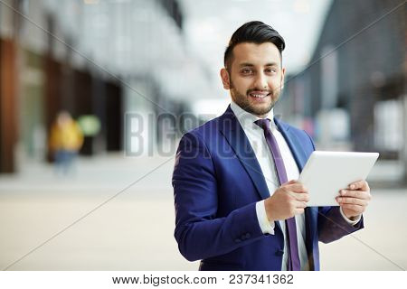 Young cheerful businessman with tablet looking at camera while analyzing online dat in airport lounge