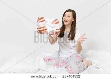 Young Brunette Woman Sitting In Bed With Red Striped Gift Box, White Sheet, Pillow, Wrapping In Blan