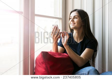 Sexy Brunette Girl Wears Blue T-shirt Holds Smartphone Sitting On Wide Windowsill With Big Red Pillo