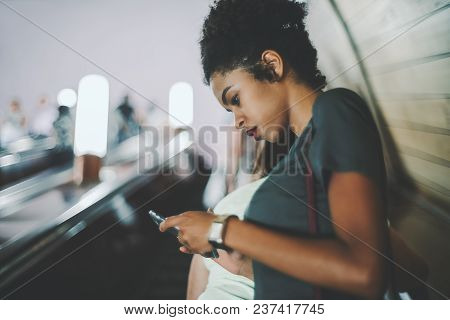 Side View Of A Curly Young Black Girl Using Her Smartphone While Standing On The Subway Escalator Mo