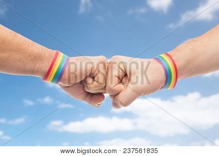 lgbt, same-sex love and homosexual relationships concept - close up of male couple hands with gay pride rainbow awareness wristbands making fist bump gesture over blue sky and clouds background