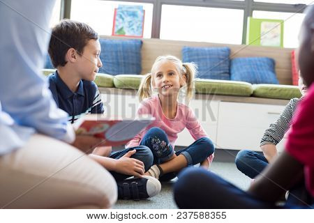 Happy smiling girl listening stories from book read by teacher at library. School children listening to fairy tale book in preschool read by woman at school. Cute girl with pigtails sitting on floor.