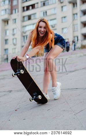 Redhead Slim Girl In Jeans Shorts And Black T Shirt Posing With Skateboard In Urban Sciene.