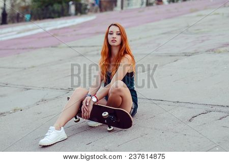 Redhead Slim Girl In Jeans Shorts And Black T Shirt Posing With Skateboard In Urban Sciene.