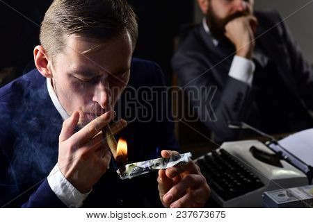 Man Lighting Cigar From Burning Banknote. Squander Concept. Businessmen In Suits Sit At Table With T