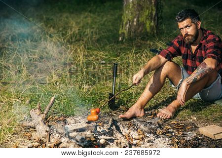 Lone Wanderer Camping In Woods. Serious Bearded Man Frying Sausages Over Fire. Barefoot Hipster Sitt