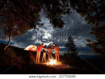 Young People Backpackers Celebrating Holiday With Beer By The Campfire Near Forest And Glowing Orang