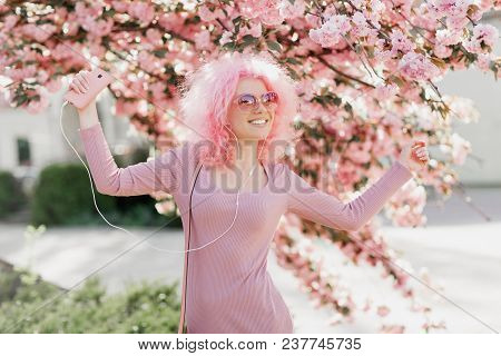 Attractive Young Woman With Curly Pink Hair And Sunglasses Near The Blossoming Spring Tree Listening