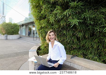 Smiling Businesswoman Working With Laptop And Papers In Open Air Near Green Plant. Concept Of Busine