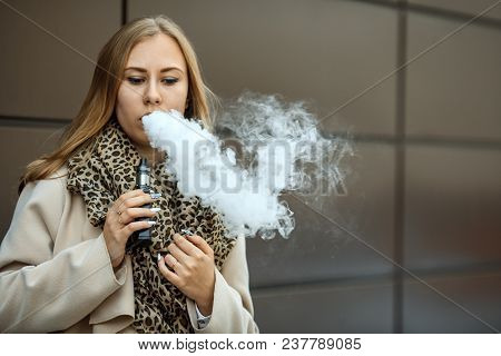 Vape Teenager. A Young Cute White Girl In Warm Scarf Is Vaping An Electronic Cigarette On The Street
