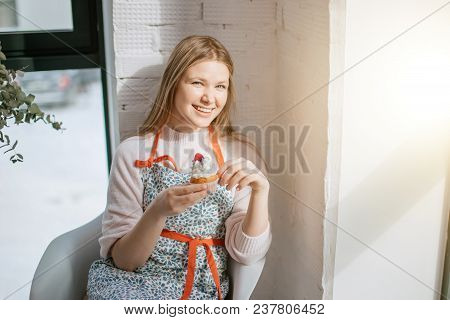 Beautiful Young Blonde Woman Preparing To Eat Capcake While Sitting Near Window, Looking At Camera.