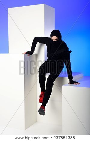 Ninja In Black Clothing With Katana Behind Getting Across On White Block Isolated On Blue