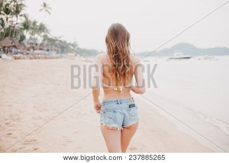 Back View Of Woman Walking On Sandy Beach In Shorts And Bikini Top