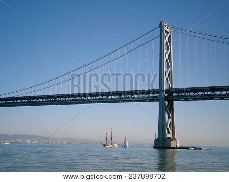 Boats Sail Under The San Francisco Side Of Bay Bridge With Oakland In The Distance On A Clear Day.