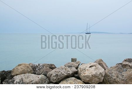 Two-masted Sailing Yacht Near Horizon And Rocks On An Overcast Day In Molinar, Mallorca, Spain.