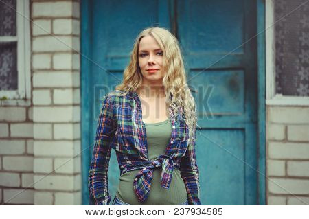 Attractive Blonde Girl On The Background Of An Old House, Dressed In Jeans And Shirts