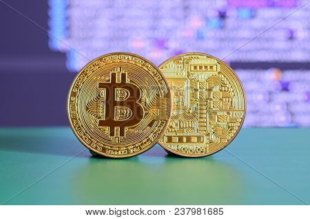 Two Gold Bitcoins Lie On The Green Surface On The Background Of The Display, Which Shows The Process