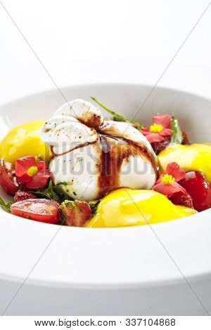 Exquisite serving burrata cheese with tomatoes and mango mousse decorated with flowers on white restaurant plate. Fresh Italian buffalo milk cheese with mozzarella, cream, arugula and pepper