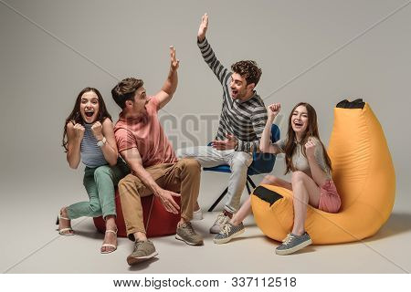 Excited Friends Giving Highfive While Sitting On Different Chairs, On Grey