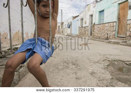 Penedo, Alagoas, Brazil - July 04, 2016: Little Kid Seated On The Entrance Gate Of His House In A Po