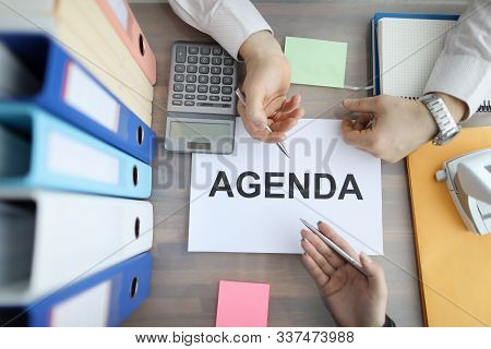Top View Of Busy Co-workers Having Heated Discussion Sitting In Office. Agenda Sign On Working Desk.
