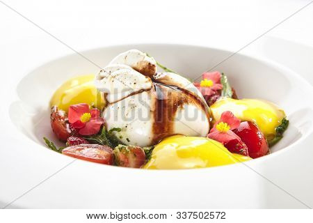 Exquisite serving burrata cheese with tomatoes and mango mousse decorated with flowers on white restaurant plate. Fresh Italian buffalo milk cheese with mozzarella, cream, arugula and pepper