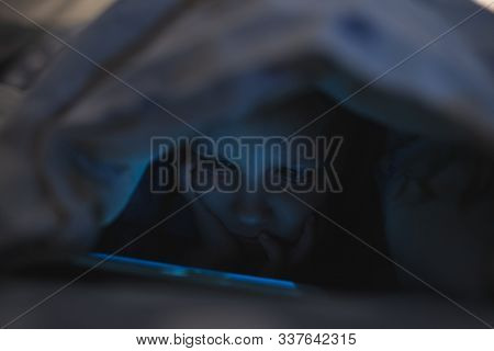 Little Girl Child Playing On Smartphone In The Evening At Home Under The Blanket When It Is Already Dark. Screen Light Reflects On Her Face