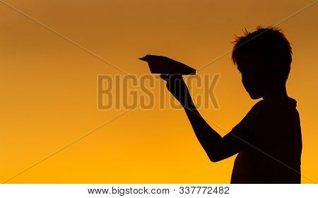 Black Silhouette Of A Boy Standing Against Orange Summer Sunset Background. Holding Small Paper Plan