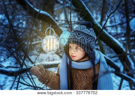 Small 4 Yaers Old Girl In Snowy Winter Forest Looks On Luminous Ball. Child In Brown Pullover, Gray 