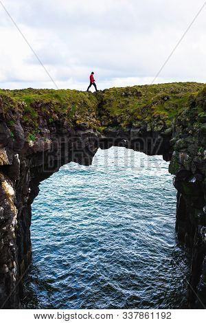 Tourist Walks Over A Natural Rock Bridge Connecting Basalt Cliffs In Arnarstapi, Iceland, With Atlan