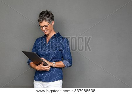 Smiling mature businesswoman wearing spectacles while using digital tablet against grey background with copy space. Beautiful middle aged woman checking email on laptop on gray wall.