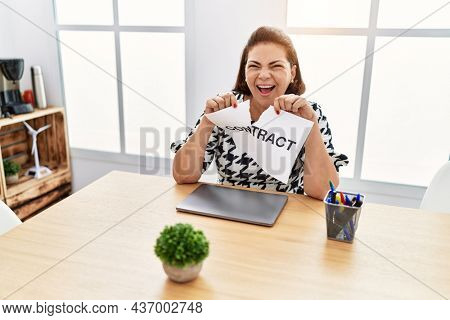 Middle age hispanic woman breaking contract paper at the office smiling and laughing hard out loud because funny crazy joke. 