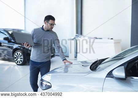 Car Salesperson With Laptop Checking Vehicle Specifications In Local Dealership Showroom.