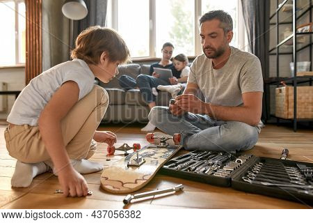 Caucasian Dad And Teenage Son Sit On Floor At Home Repair Skateboard With Toolset In Box. Young Fath