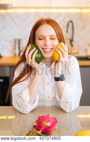 Vertical Portrait Of Cheerful Attractive Redhead Young Woman Holding In Hands Mango And Avocado Sitt